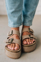 Beige sandals with buckles worn with light blue jeans on a neutral background