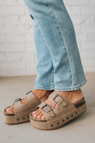 Person wearing beige platform sandals with light blue jeans against a white brick wall.