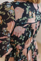 Close-up of a floral dress with a black background, large pink roses, green leaves, and small blue and orange flowers. The dress has a gathered waist and short puff sleeves.