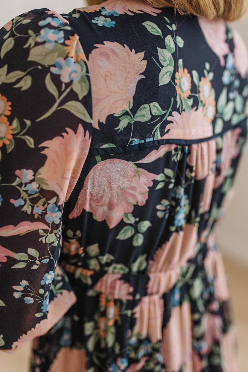 Close-up of a floral dress with a black background, large pink roses, green leaves, and small blue and orange flowers. The dress has a gathered waist and short puff sleeves.