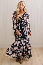A woman modeling a long-sleeve maxi dress with a dark navy blue base and a pink floral pattern, featuring a matching navy blue tie belt at the waist, set against a light wood-paneled wall.
