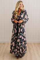 A woman modeling a long-sleeve maxi wrap dress with a black base and a pink floral pattern, standing in front of a white paneled wall and light wood floor.
