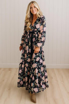 A woman modeling a long-sleeve maxi dress with a black base and a pink floral pattern, standing in front of a white paneled wall and light wood floor.
