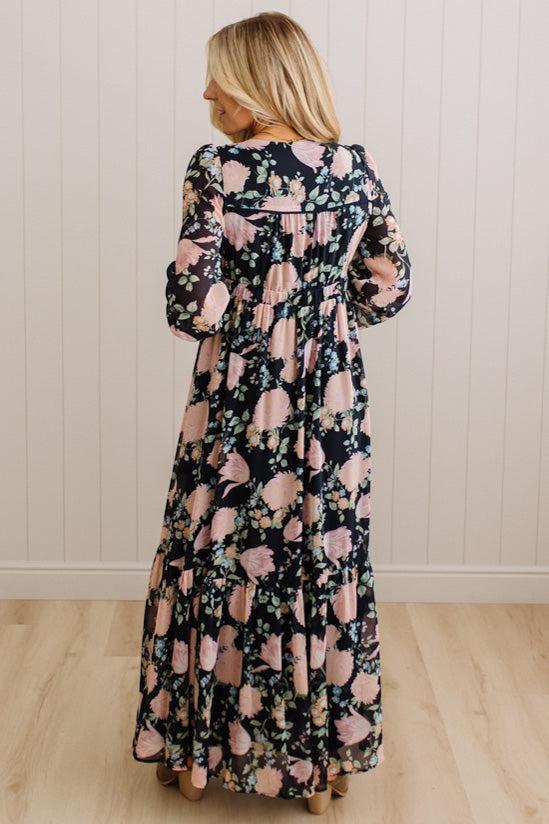 A woman wearing a long-sleeved maxi dress with a pink and green floral pattern on a dark navy background, shown from the back against a white paneled wall and light wood floor.