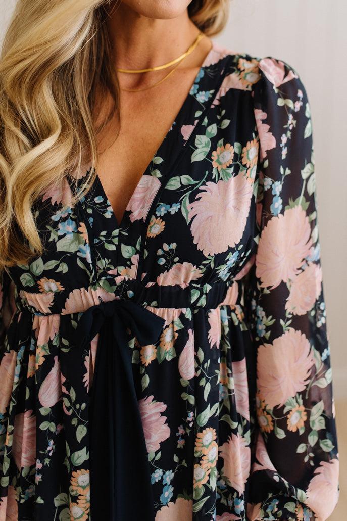A close-up of a woman wearing a long-sleeve wrap dress with a black base and a large pink, peach, and green floral print. The dress features a deep V-neckline, a gathered waist with a matching fabric tie belt, and puffed sleeves.