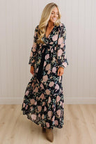 A woman wearing a long-sleeve maxi dress with a black base and a pink floral pattern, standing in a room with light wood floors and white paneled walls.