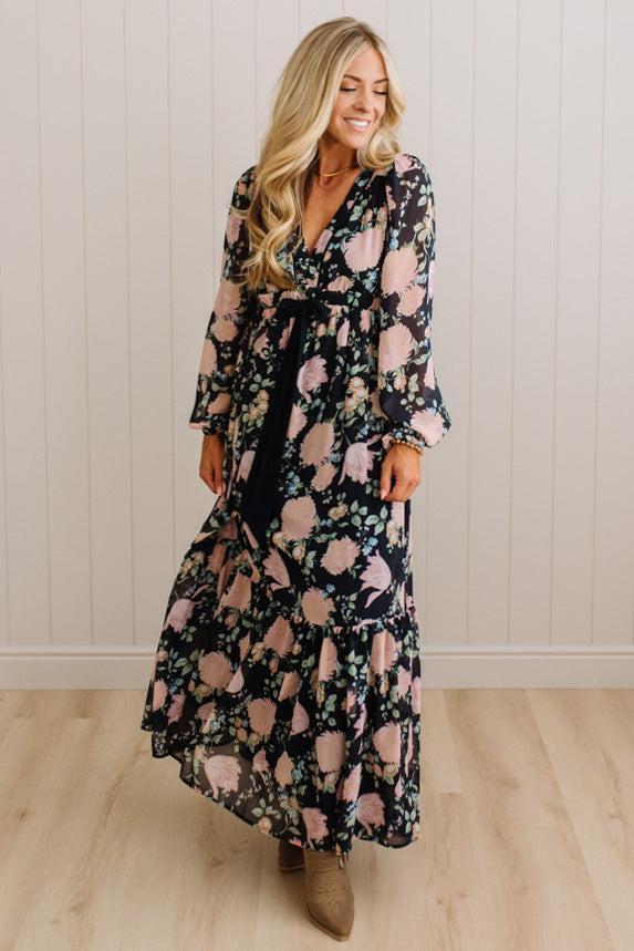 A woman wearing a long-sleeve maxi dress with a black base and a pink floral pattern, standing in a room with light wood floors and white paneled walls.