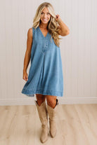 A woman modeling a sleeveless light blue denim dress with a V-neck and button placket, paired with beige cowboy boots, standing in front of a white paneled wall and light wood floor.