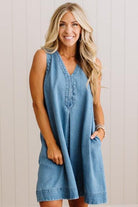 A woman wearing a sleeveless light blue denim dress with a V-neck and button placket, standing against a light beige paneled wall.