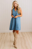 A woman modeling a sleeveless blue denim dress with a V-neck and button placket, paired with beige cowboy boots, standing in front of a white paneled wall and light wood floor.