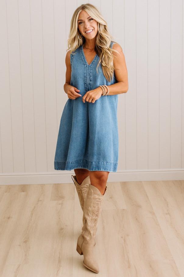 A woman modeling a sleeveless blue denim dress with a V-neck and button placket, paired with beige cowboy boots, standing in front of a white paneled wall and light wood floor.