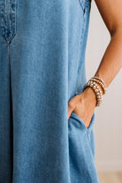 Close-up of a person wearing a blue denim jumpsuit, with their hand in the pocket and stacked pearl beaded bracelets on one wrist, set against a neutral light beige background.