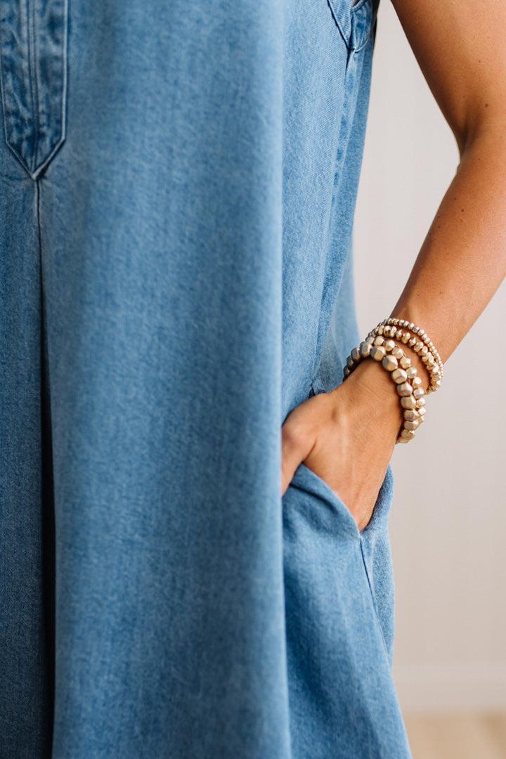 Close-up of a person wearing a blue denim jumpsuit, with their hand in the pocket and stacked pearl beaded bracelets on one wrist, set against a neutral light beige background.