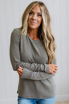 A woman with blonde hair wearing a long-sleeve crewneck top with thin horizontal gray and white stripes, paired with blue jeans, standing against a white paneled wall.