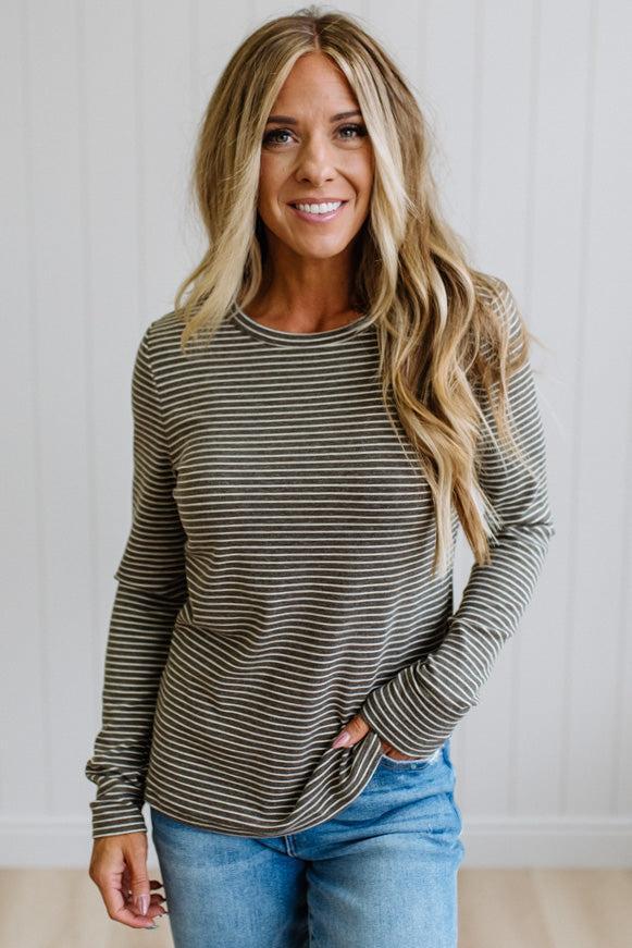 A woman modeling a long-sleeve striped knit top and blue jeans in a bright room with white paneled walls and light wood flooring.