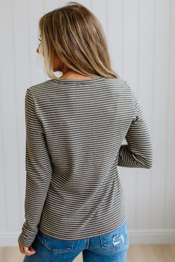 A woman shown from the back wearing a long-sleeve shirt with thin horizontal gray and white stripes, paired with blue jeans, against a white paneled wall.