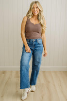 Woman wearing a brown tank top and blue jeans standing against a light wooden paneled wall.