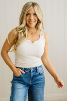 Woman wearing a white tank top and blue jeans against a light wooden paneled wall.