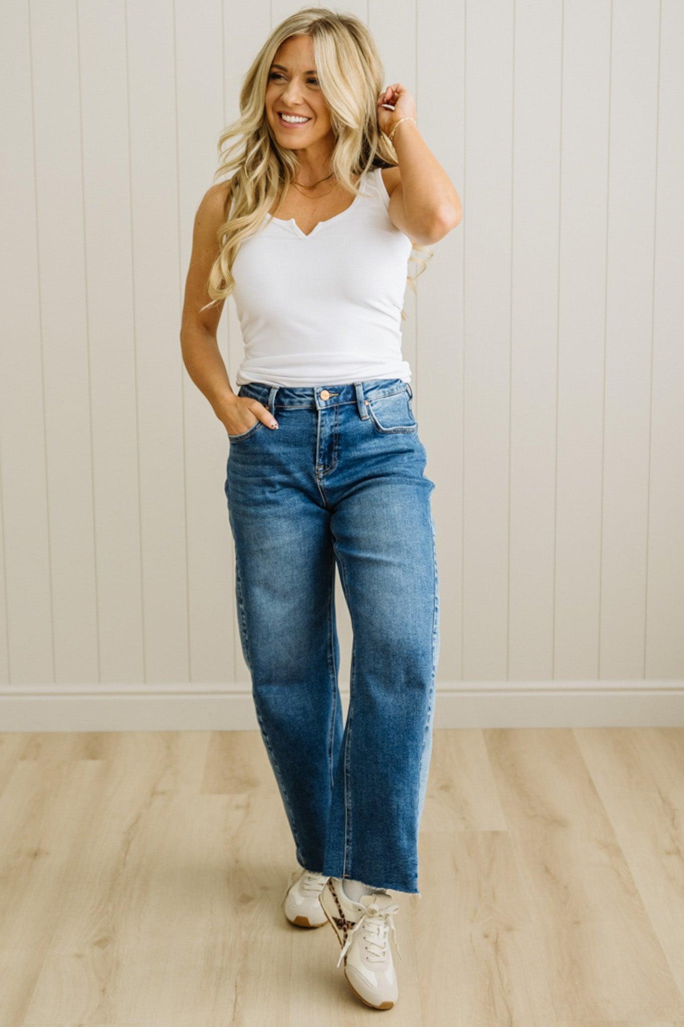 Woman wearing a white tank top and blue jeans standing against a light wooden paneled wall.