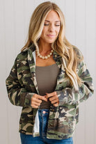 A woman wearing a green and brown camouflage button-up shirt jacket over a taupe tank top and blue jeans, standing against a white paneled wall.