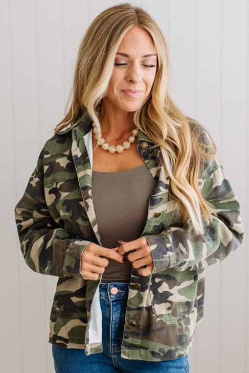 A woman wearing a green and brown camouflage button-up shirt jacket over a taupe tank top and blue jeans, standing against a white paneled wall.
