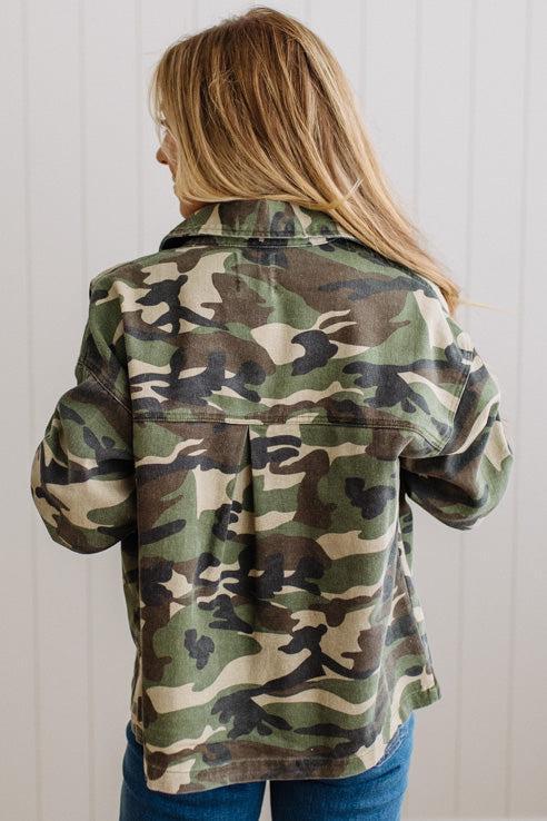 A woman viewed from behind wearing a camouflage jacket over a light top and blue jeans, standing against a white paneled wall.