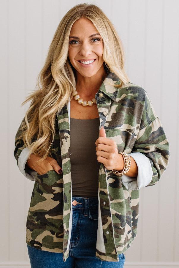 A smiling blonde woman wearing a green, brown, and beige camouflage button-up shirt jacket over a dark brown tank top and blue jeans, standing against a white paneled wall.