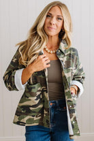 A woman wearing a green and brown camouflage flannel button-up shirt over a taupe tank top and blue jeans, standing against a white paneled wall.