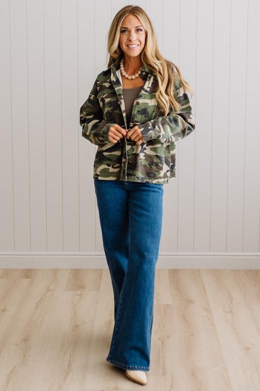 A woman modeling a camouflage button-up jacket over a gray top and blue flare jeans, standing against a white paneled wall on a light wood floor.
