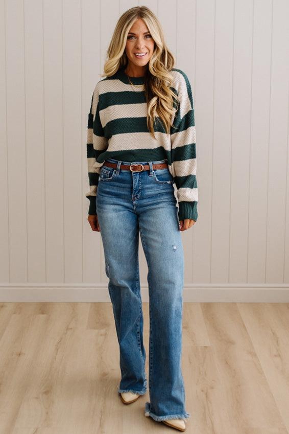 A woman modeling a dark green and cream striped knit sweater paired with blue flare jeans, a brown belt, and beige pointed-toe flats, standing against a white paneled wall and light wood floor.