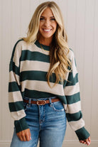 A smiling blonde woman wearing a dark green and cream waffle-knit sweater, blue jeans, and a brown leather belt, standing against a light beige paneled wall.