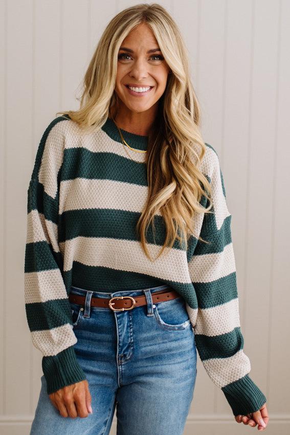 A smiling blonde woman wearing a dark green and cream waffle-knit sweater, blue jeans, and a brown leather belt, standing against a light beige paneled wall.