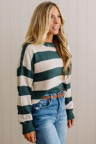 A woman wearing a dark green and cream horizontally striped knit sweater, tucked into blue jeans with a brown belt, standing against a light beige paneled wall.
