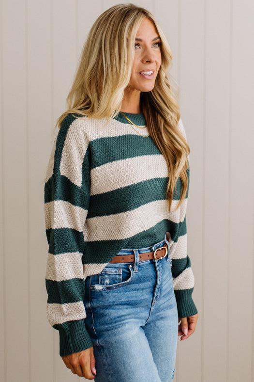 A woman wearing a dark green and cream horizontally striped knit sweater, tucked into blue jeans with a brown belt, standing against a light beige paneled wall.