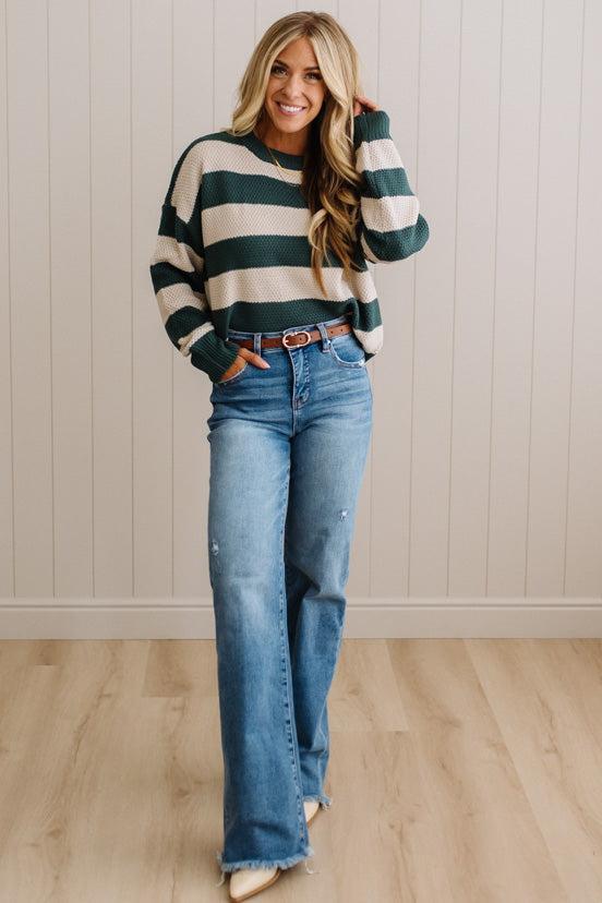 A woman modeling a dark green and cream striped knit sweater with blue flare jeans, standing against a light wood-paneled wall.