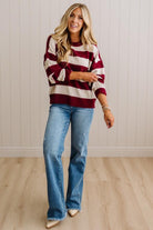 A woman modeling a long-sleeve knit sweater with bold horizontal burgundy and cream stripes, paired with light-wash blue flare jeans and beige pointed-toe heels, standing against a white paneled wall and light wood floor.