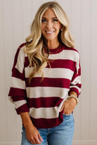 A smiling blonde woman wearing a maroon and cream horizontally striped knit sweater with blue jeans, standing against a light beige paneled wall.