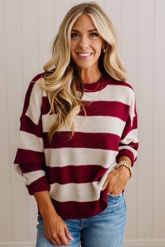 A smiling blonde woman wearing a maroon and cream horizontally striped knit sweater with blue jeans, standing against a light beige paneled wall.