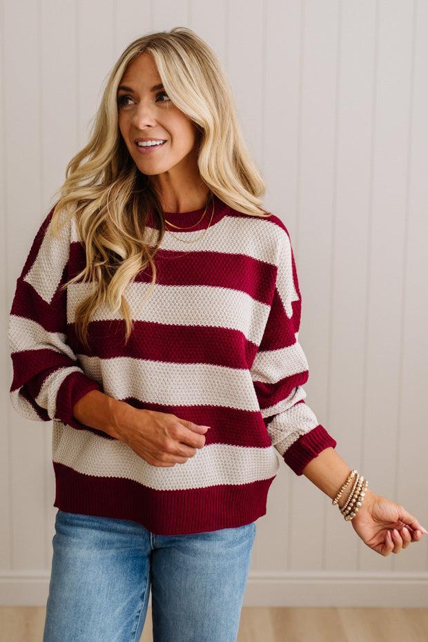 A woman wearing a knit sweater with horizontal burgundy and cream stripes, paired with blue jeans, standing against a white paneled wall.
