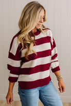 A woman wearing a knit sweater with horizontal burgundy and cream stripes, paired with light blue jeans, standing against a white paneled wall.