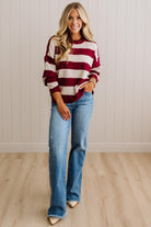A woman modeling a red and cream striped knit sweater with light-wash blue jeans, standing in front of a white paneled wall and light wood floor.