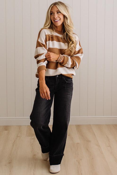 A woman modeling a brown and cream striped knit sweater, paired with dark wash jeans and white shoes, standing against a white paneled wall and light wood floor.