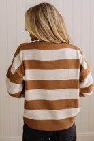 A woman viewed from behind wearing a waffle-knit sweater with horizontal stripes in warm brown and cream, paired with dark jeans against a white paneled wall.