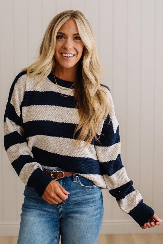 A woman with long blonde hair wearing a navy and cream horizontally striped knit sweater and blue jeans, standing against a white paneled wall.