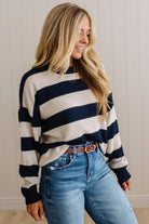 A woman wearing a navy and cream striped knit sweater with blue jeans, standing against a light beige paneled wall.