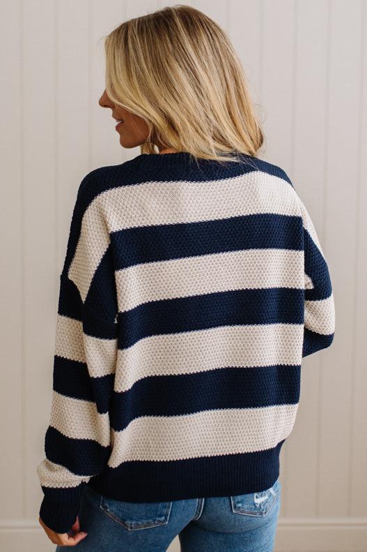 A woman shown from the back wearing a navy blue and cream waffle-knit sweater with horizontal stripes, paired with blue jeans, against a white paneled wall.