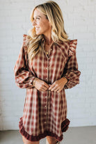 Woman wearing a plaid dress with ruffled sleeves and hem against a white brick wall.