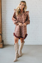 Woman wearing a plaid dress with ruffled details and beige knee-high boots against a white brick wall.