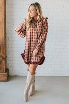 Woman wearing a plaid dress with ruffled details and beige knee-high boots against a white brick wall.