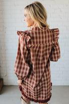 Woman wearing a plaid dress with ruffled sleeves and bottom in front of a white brick wall.
